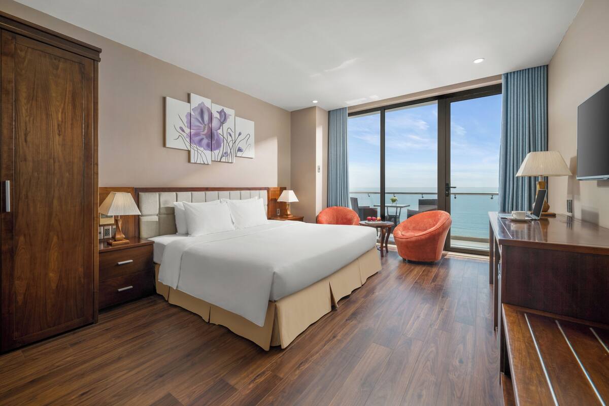 A hotel room with a large bed, a desk, a chair, a TV, and a view of the sea.