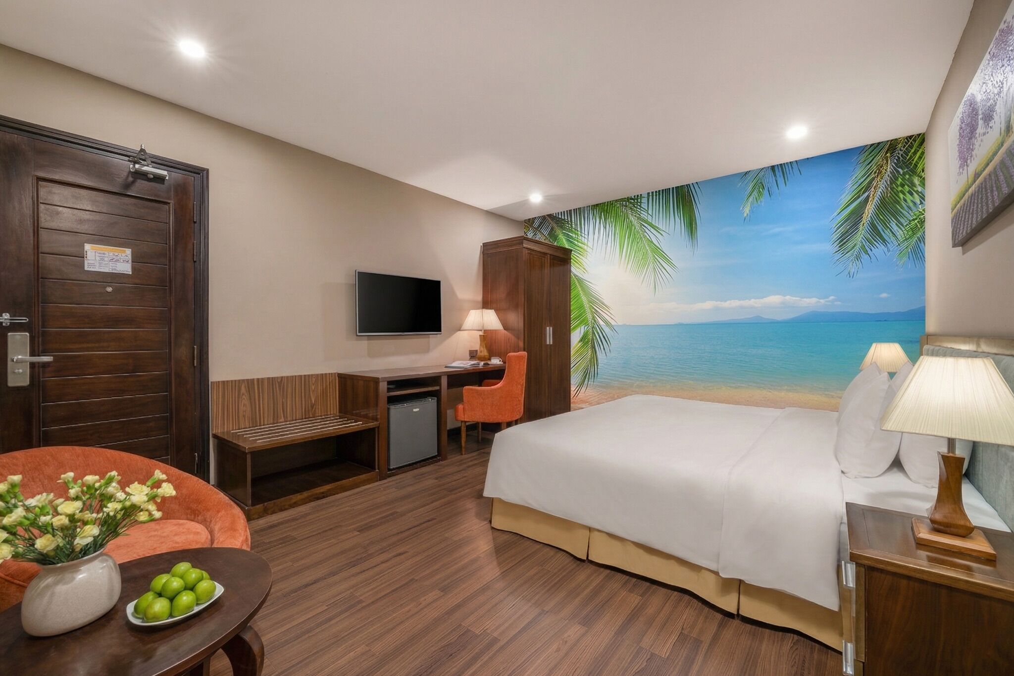 A hotel room with a bed, desk, chair, TV, and a beach mural on the wall.