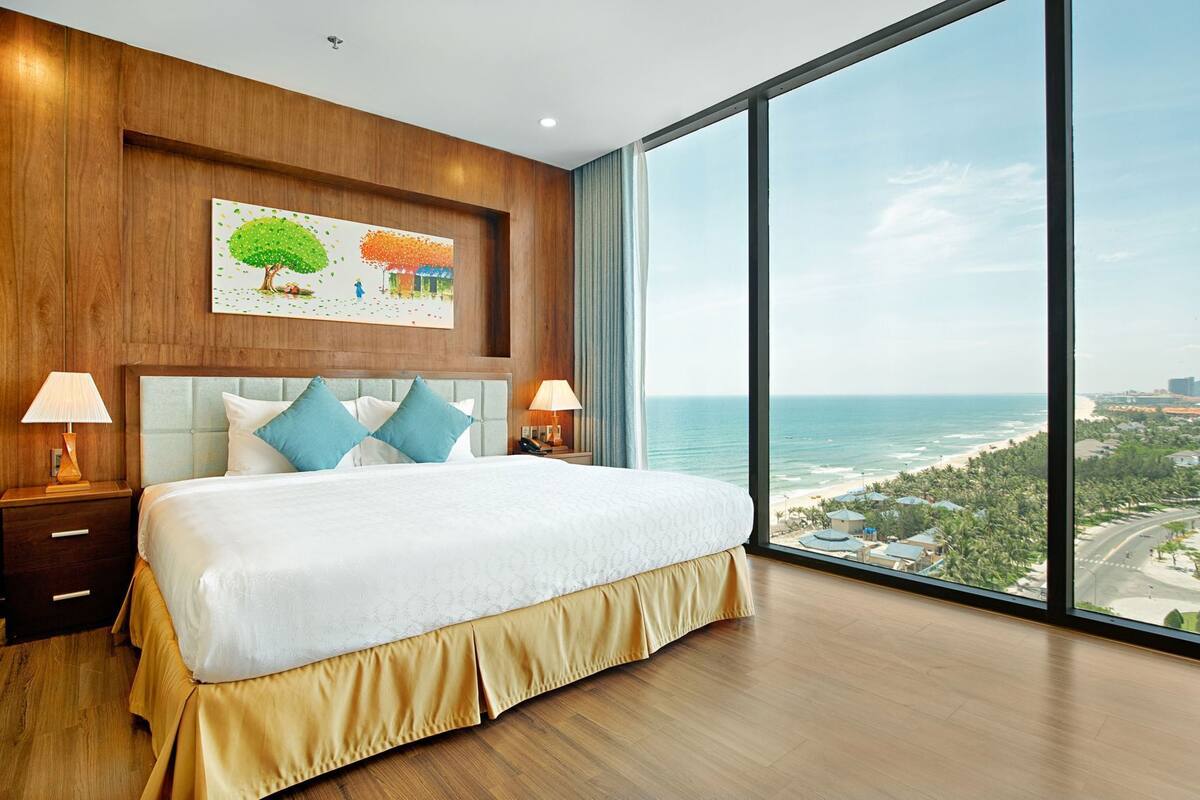 A bedroom with a large bed, two bedside tables with lamps, a wooden headboard, a painting, and a large window with a view of the ocean and cityscape.