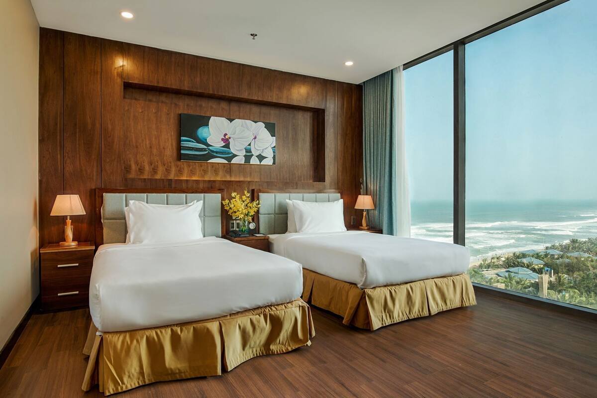 A hotel room with two beds, wooden paneling, a large window with a view of the ocean, and a painting of white flowers.