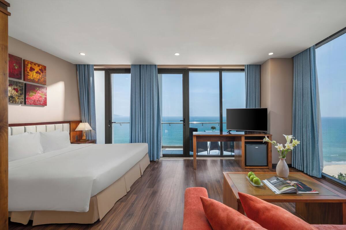 A hotel room with a bed, a TV, a desk, and a view of the ocean.