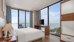 A hotel room with a bed, a desk with a TV, a wardrobe, and a large window with a view of the beach. - Yarra Ocean Suites Danang (Da Nang)