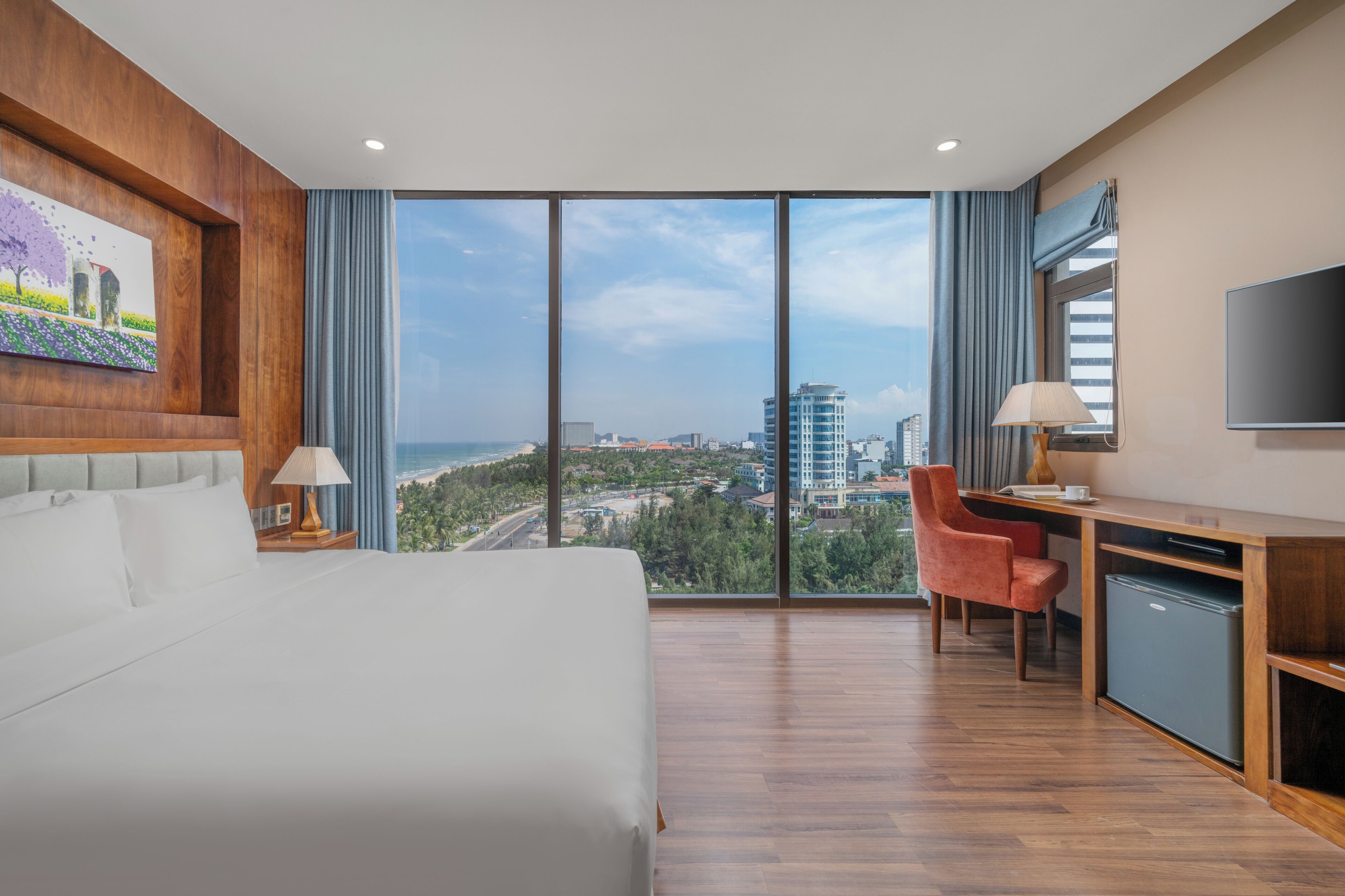 Ein Hotelzimmer mit Bett, Schreibtisch, Stuhl, Fernseher und einem großen Fenster mit Blick auf die Stadt.