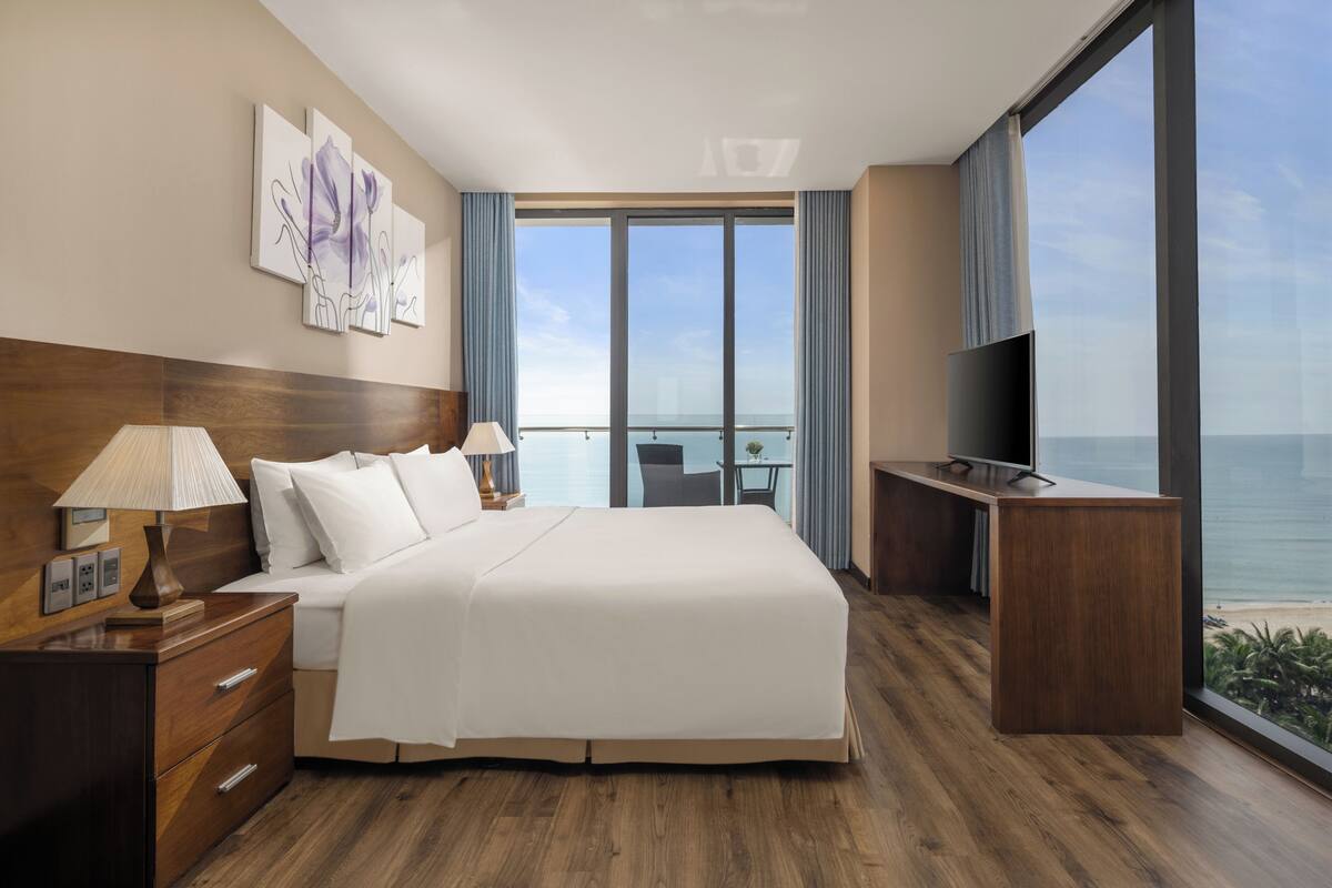 A hotel room with a large bed, wooden floor, a TV, a desk, and a balcony with a view of the sea.