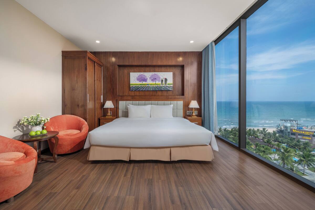 A bedroom with a large bed, two side tables, a chair, a painting, and a view of the ocean.