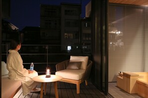 Un balcon avec une personne assise sur un canapé, une table avec une bouteille et une bougie, et un banc en bois.