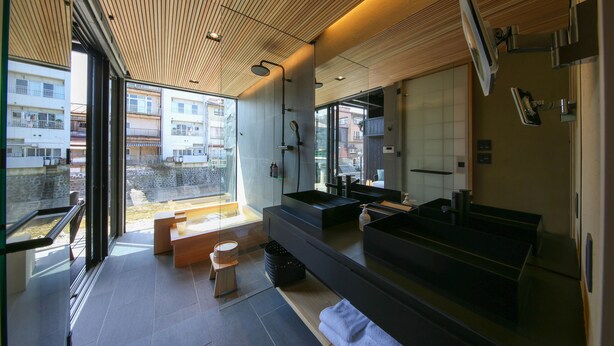 Une salle de bain moderne avec un grand miroir, une baignoire et un lavabo. On y trouve un banc et un petit tabouret. La pièce est dotée d’un plafond en bois et de grandes fenêtres.