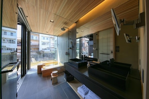 A modern bathroom with a large mirror, a bathtub, and a sink. There is a bench and a small stool. The room has a wooden ceiling and large windows.