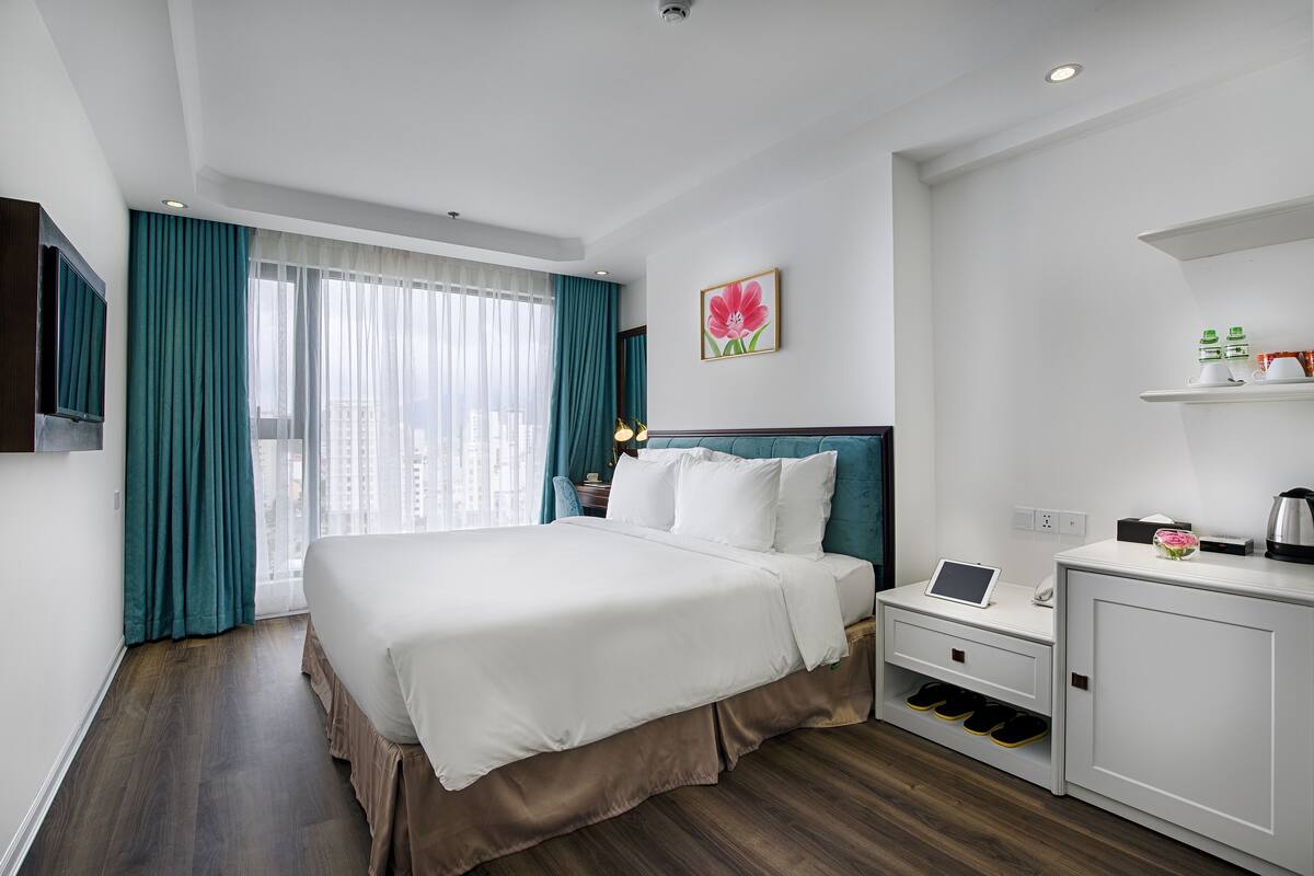 Une chambre d’hôtel avec un grand lit, un bureau avec un ordinateur portable, une armoire à chaussures et une fenêtre avec des rideaux.