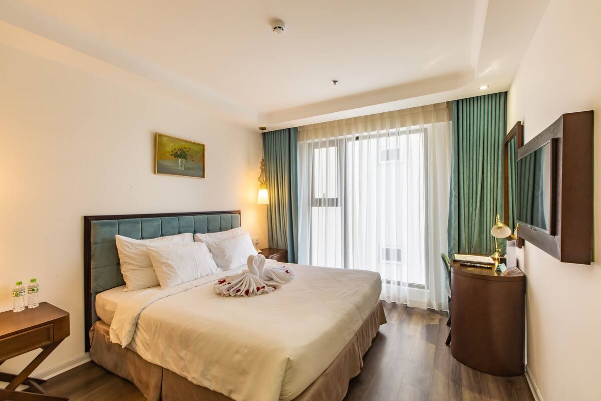 Une chambre d’hôtel avec un grand lit, une table de chevet avec une lampe, un tableau au mur et une fenêtre avec des rideaux.