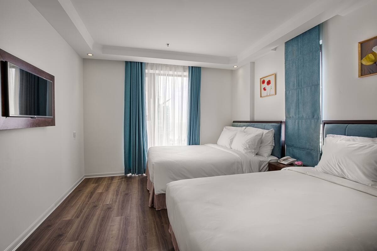 Une chambre d’hôtel avec deux lits, une grande fenêtre avec des rideaux bleus, un plancher en bois et un téléviseur fixé au mur.