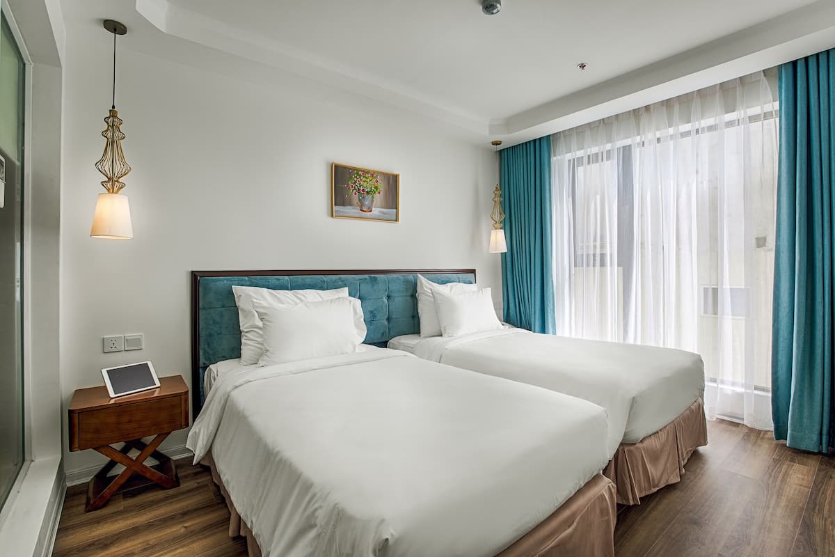 Une chambre d’hôtel avec deux lits, une table de chevet avec une tablette, un tableau au mur et une fenêtre avec des rideaux.