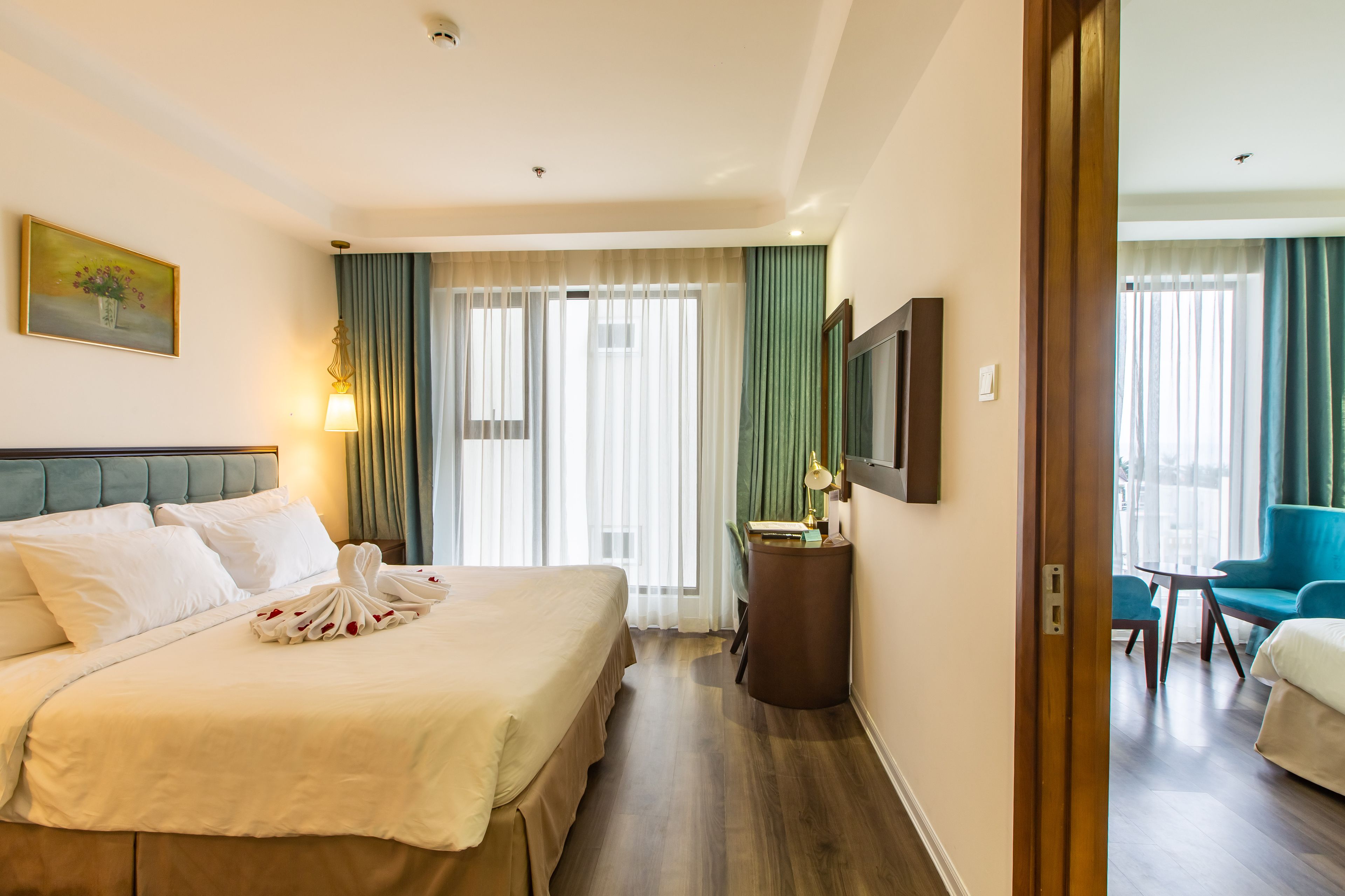 A hotel room with a large bed, a desk with a lamp, a chair, and a view of the outside through the windows.
