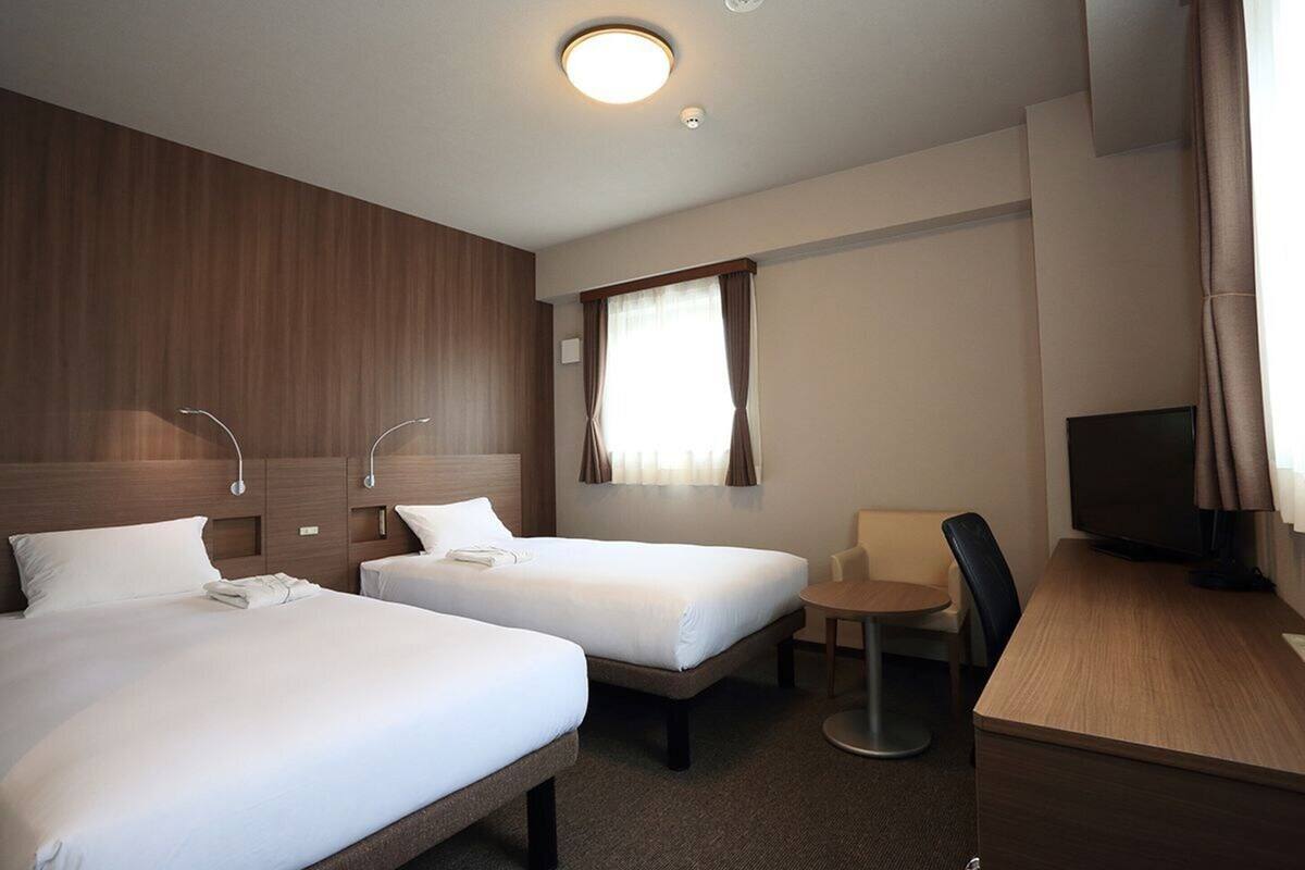 Une chambre d’hôtel avec deux lits simples, un bureau, une chaise, une télévision et une fenêtre avec des rideaux.