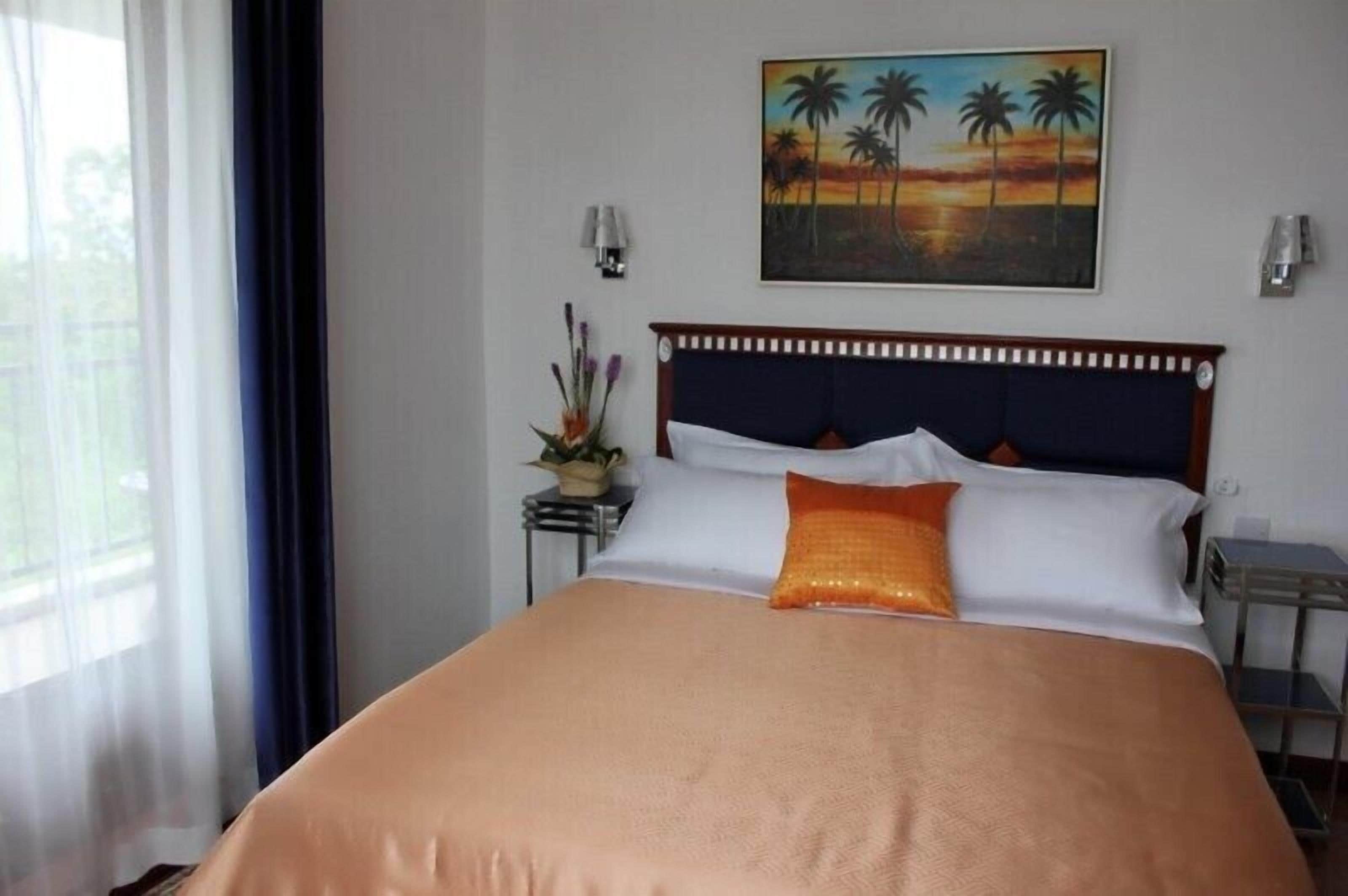 Une chambre d’hôtel avec un lit, un tableau représentant des palmiers, une table de chevet et une lampe.