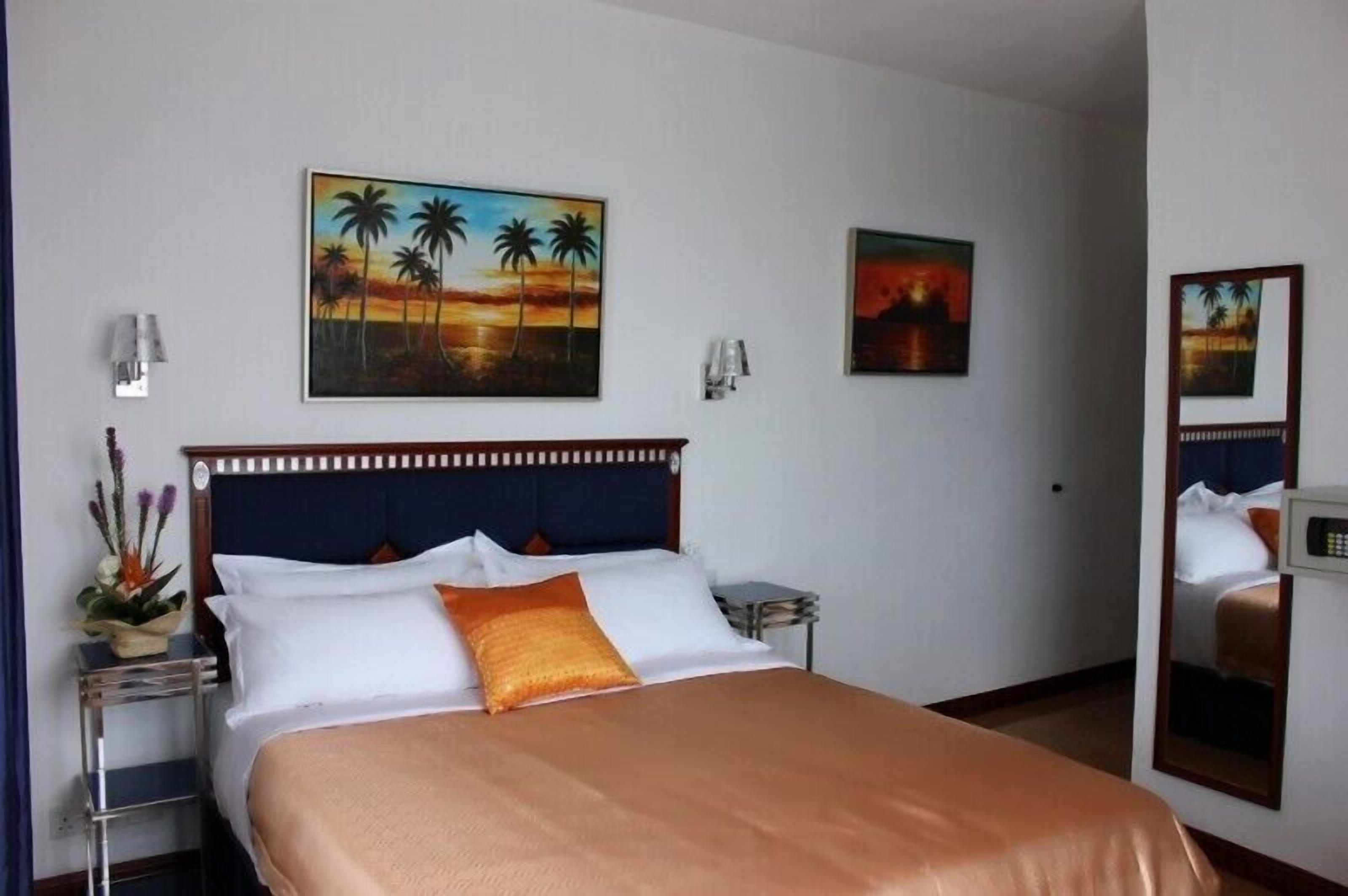 Una camera d'albergo con un letto, un comodino, una lampada, un quadro con palme e uno specchio.