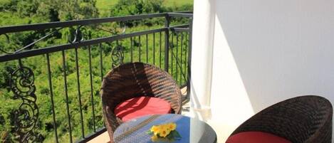 A balcony with wicker furniture, a small table with a yellow flower, and a view of a green landscape.