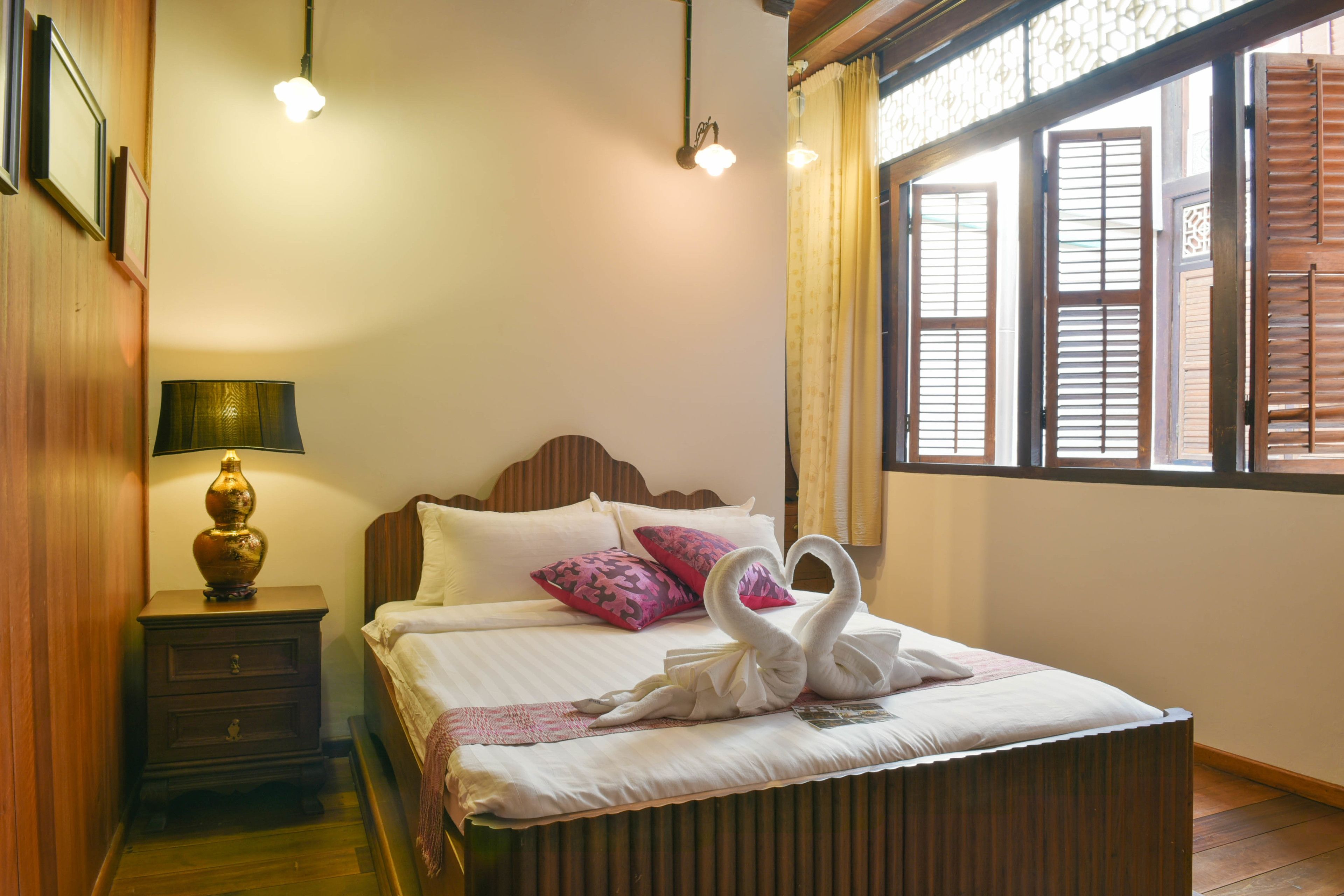 A neatly made bed with a swan-shaped pillow arrangement, a bedside lamp, and a window with wooden shutters.