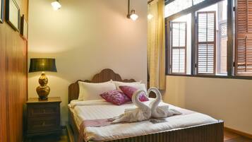 A neatly made bed with a swan-shaped pillow arrangement, a bedside lamp, and a window with wooden shutters.