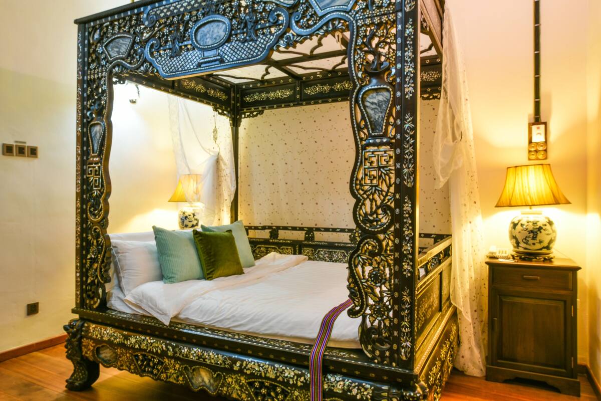 A four-poster bed with ornate carvings, a bedside table with a lamp, and a small cabinet.