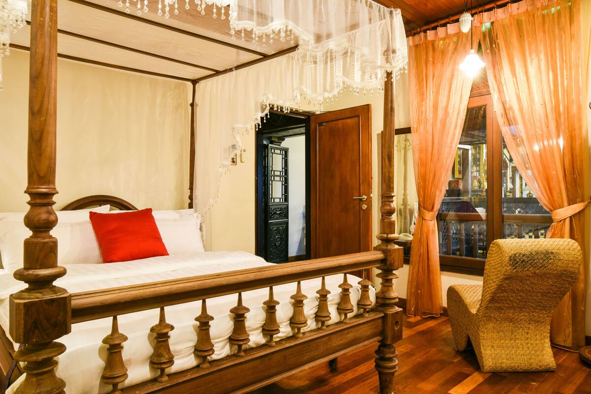 A four-poster bed with a wooden headboard, a red pillow, and a chair with a woven seat.