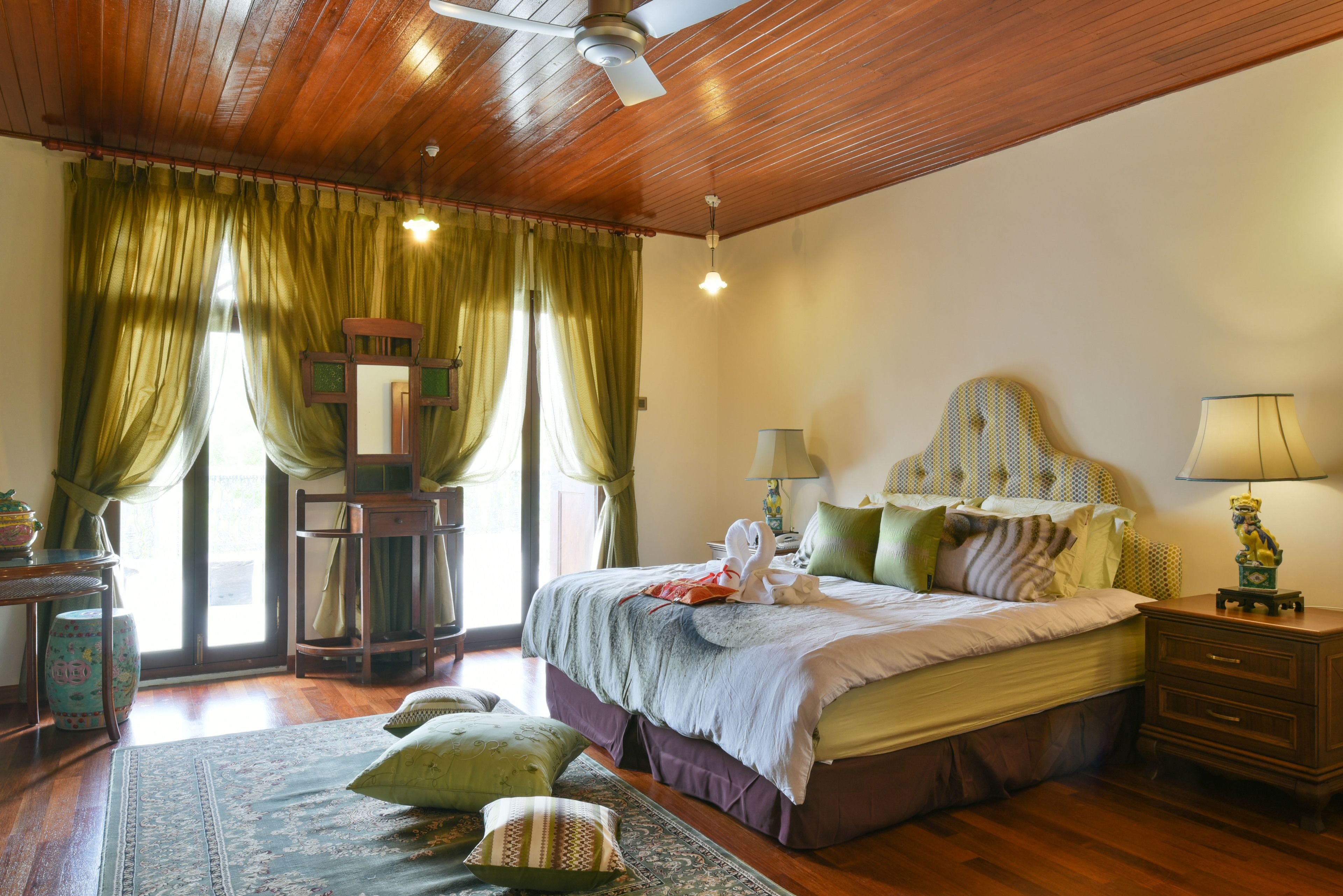 A bedroom with a wooden ceiling, a bed with green and white bedding, a nightstand with a lamp, and a window with green curtains.