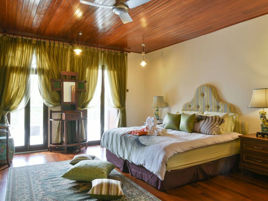 A bedroom with a wooden ceiling, a bed with green and white bedding, a nightstand with a lamp, and a window with green curtains.