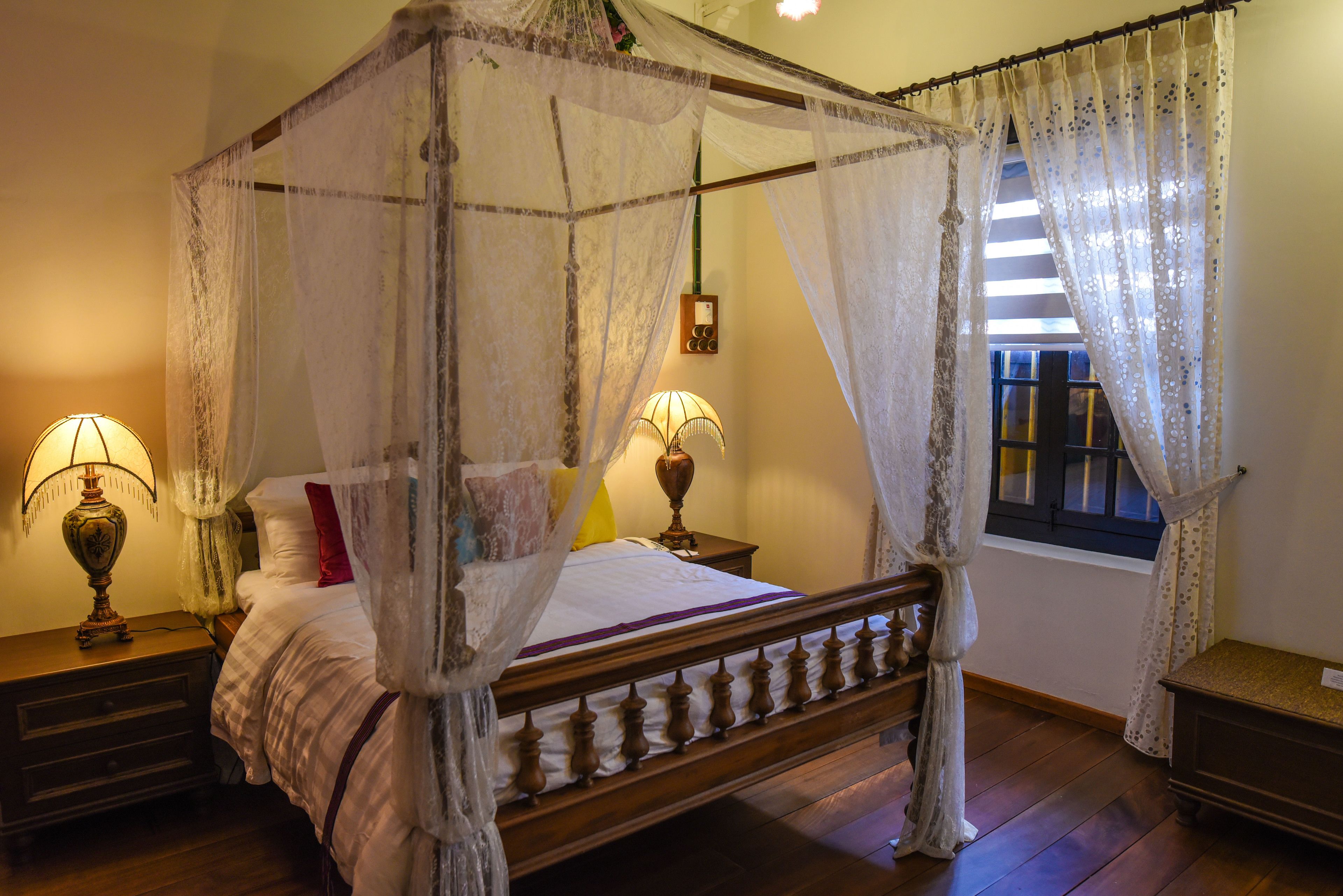 A four-poster canopy bed with a wooden frame, two bedside tables with lamps, a window with curtains, and a bedside table with a clock.