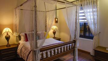 A four-poster canopy bed with a wooden frame, two bedside tables with lamps, a window with curtains, and a bedside table with a clock.