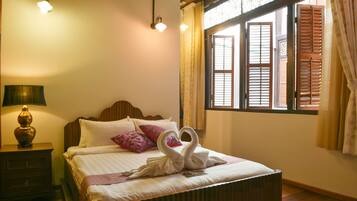 A neatly made bed with a swan-shaped towel arrangement, a bedside lamp, and a window with wooden shutters.