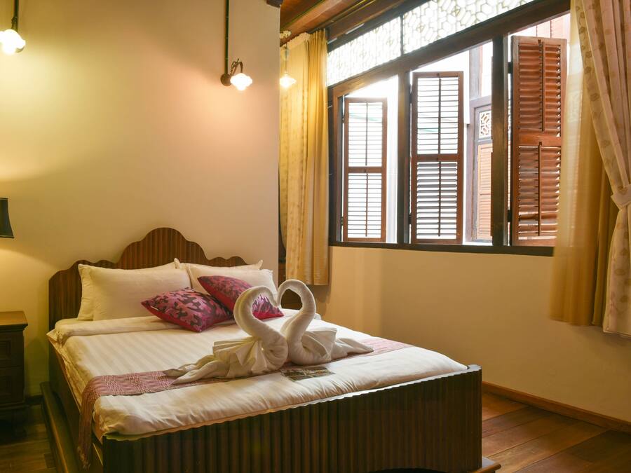 A neatly made bed with a swan-shaped towel arrangement, a bedside lamp, and a window with wooden shutters.