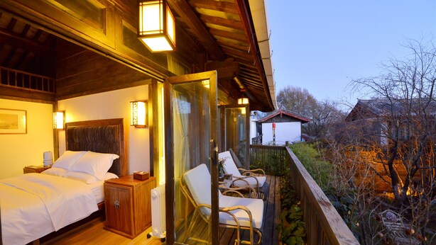 A bedroom with two beds, a wooden headboard, bedside tables, and a glass door leading to a balcony with wicker chairs and a view of the surrounding area.