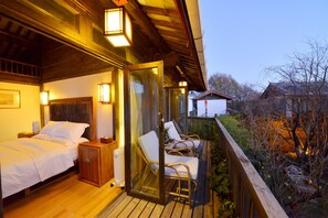 Une chambre avec deux lits, une tête de lit en bois, des tables de chevet et une porte vitrée donnant sur un balcon équipé de chaises en osier et offrant une vue sur les environs.