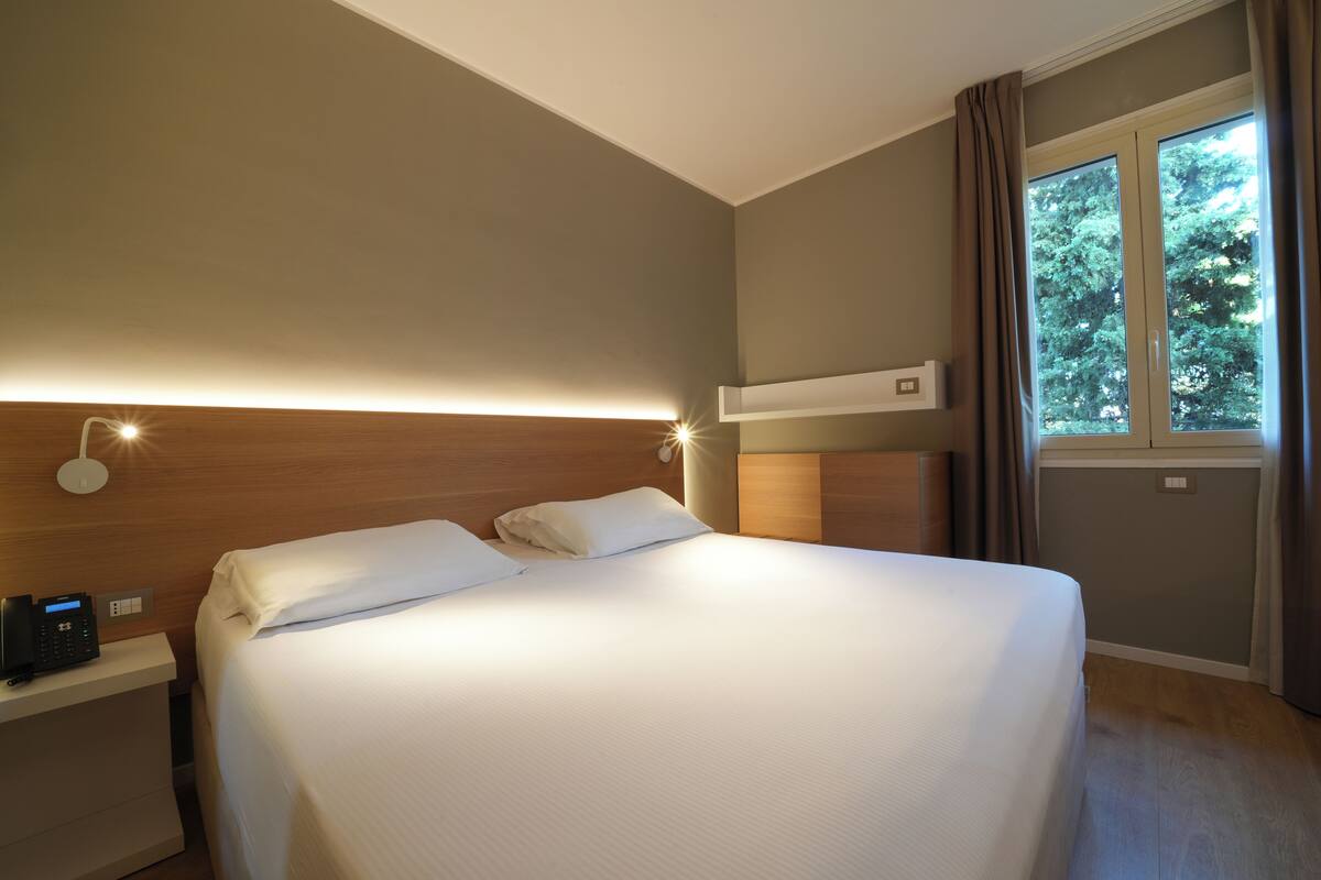 A hotel room with a large bed, bedside tables, a control panel, and a window with curtains.