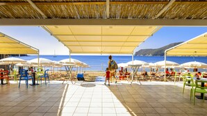 An outdoor seating area with tables and chairs under umbrellas, overlooking a beach with a clear view of the sea and distant hills. - TH Lacona (Capoliveri)