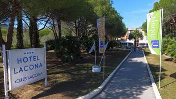 A pathway leading to a hotel named 'HOTEL LACONA CLUB LACONA' with flags and trees in the background.