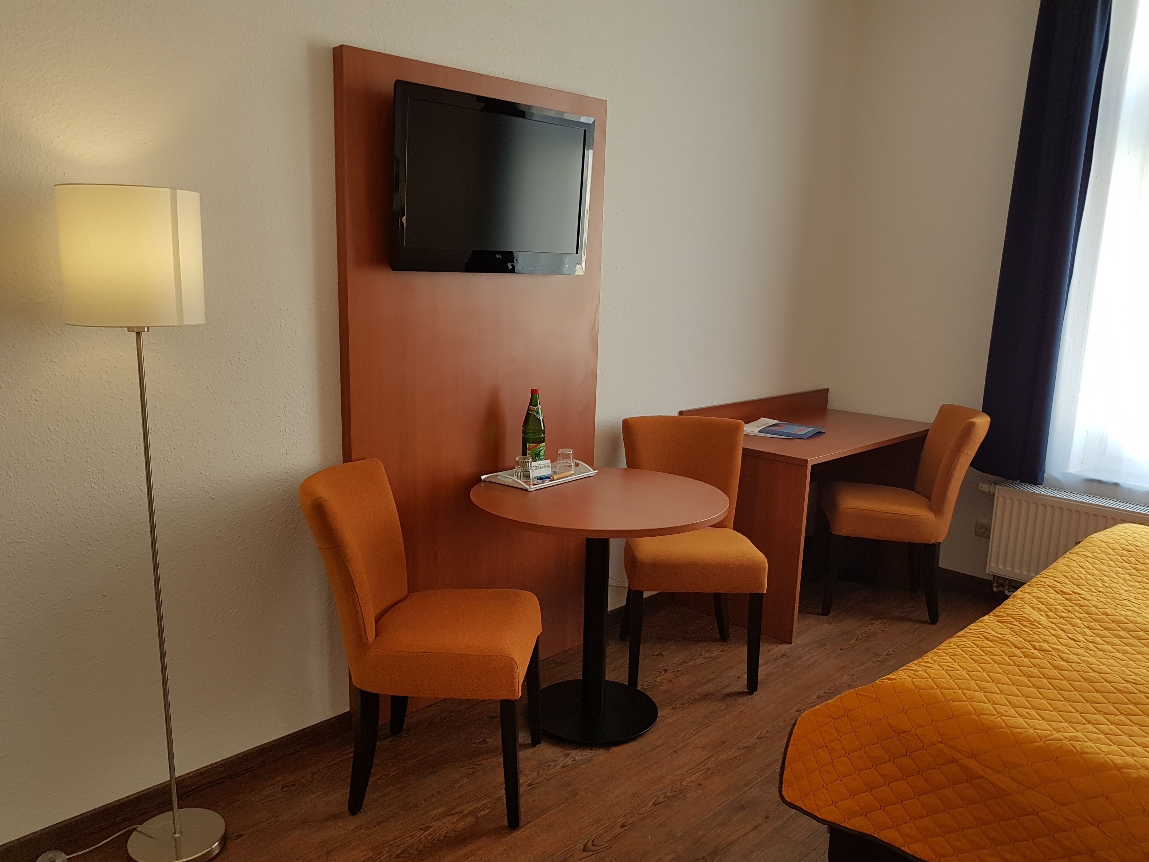 Habitación de hotel con televisión montada en la pared, una mesa redonda con dos sillas, una cama con colcha amarilla y una lámpara de pie.
