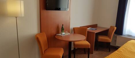 Habitación de hotel con televisión montada en la pared, una mesa redonda con dos sillas, una cama con colcha amarilla y una lámpara de pie.