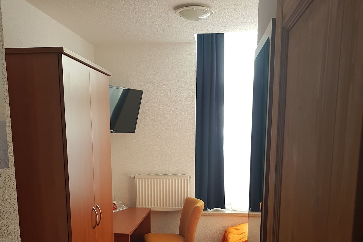 Quarto de hotel pequeno com guarda-roupa de madeira, escrivaninha, uma cama individual e janela com cortinas.