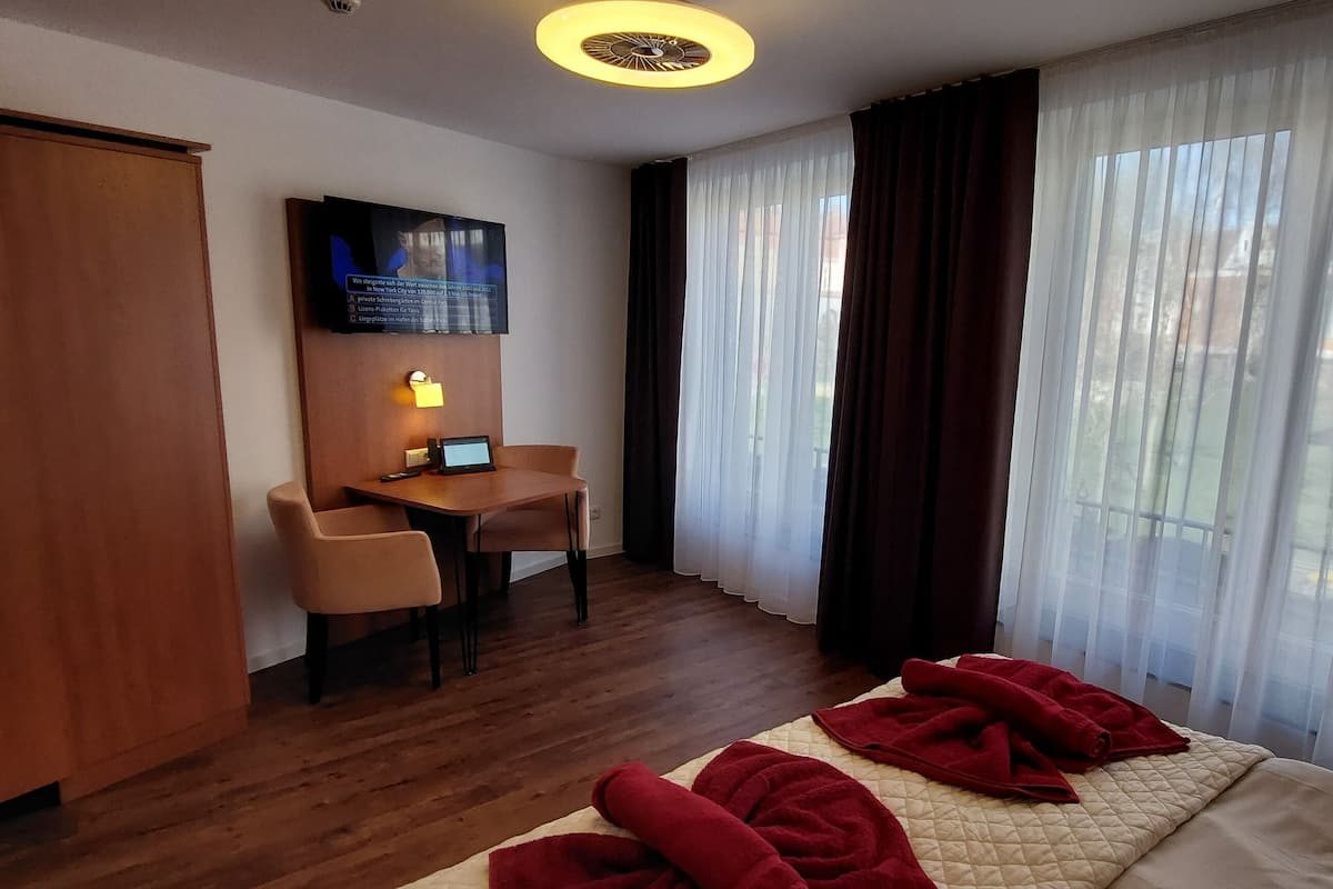 Quarto de hotel com cama, escrivaninha, cadeira, televisão e janela com cortinas.
