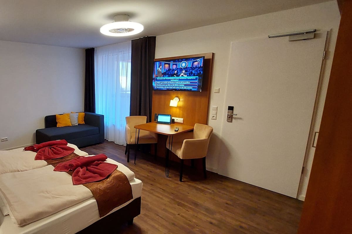 Quarto de hotel com cama, secretária, cadeira, televisão e porta.
