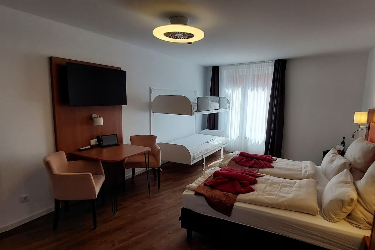 Quarto de hotel com cama, escrivaninha, cadeira, televisão e janela com cortinas.