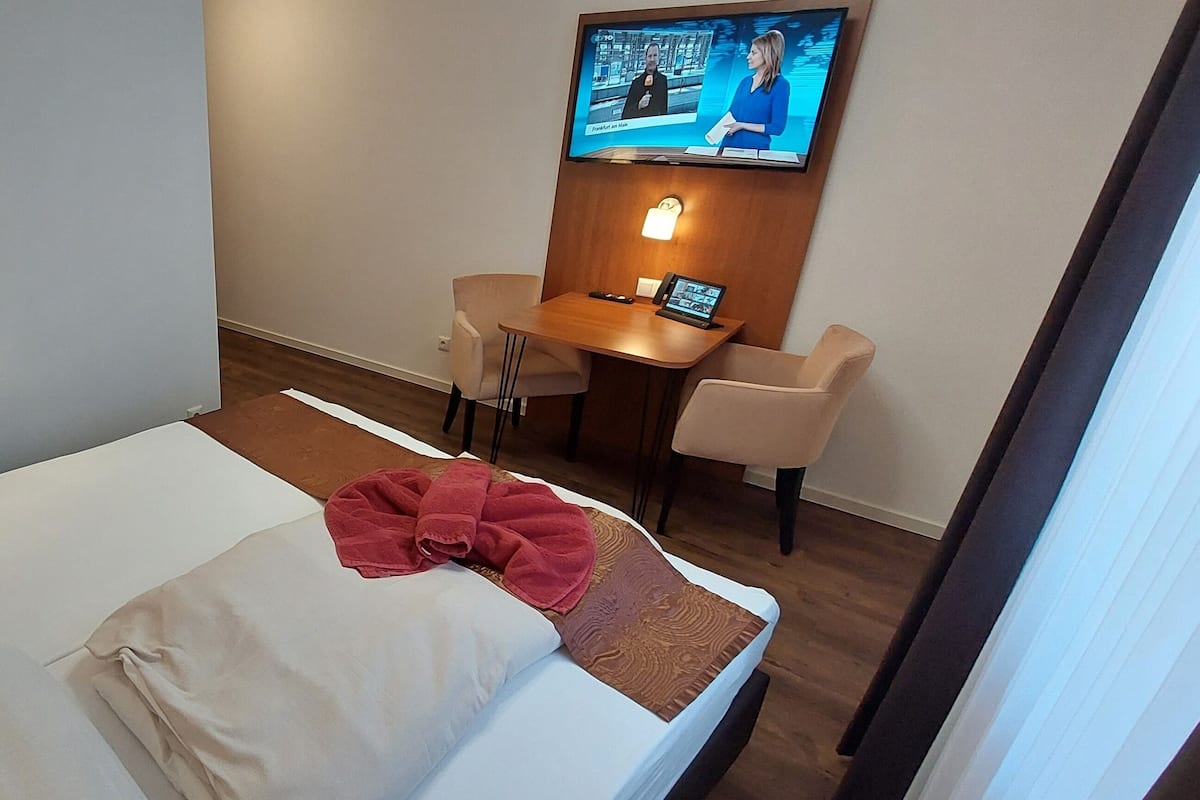Quarto de hotel com cama, escrivaninha e televisão fixada na parede.