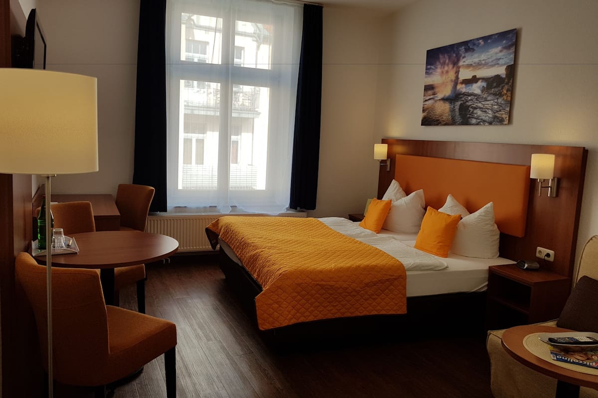 Quarto de hotel com cama, escrivaninha, cadeira, mesa e um quadro na parede.