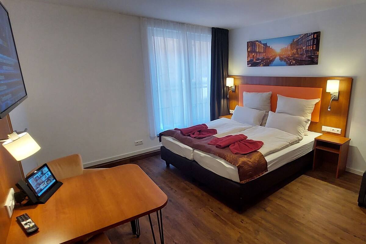 Quarto de hotel com cama, mesas de cabeceira, escrivaninha, televisão e um quadro na parede.