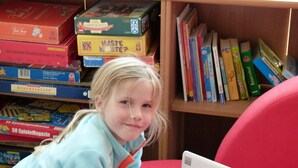 Een kind leest een boek in een bibliotheek met boekenplanken vol boeken en lesmateriaal.
