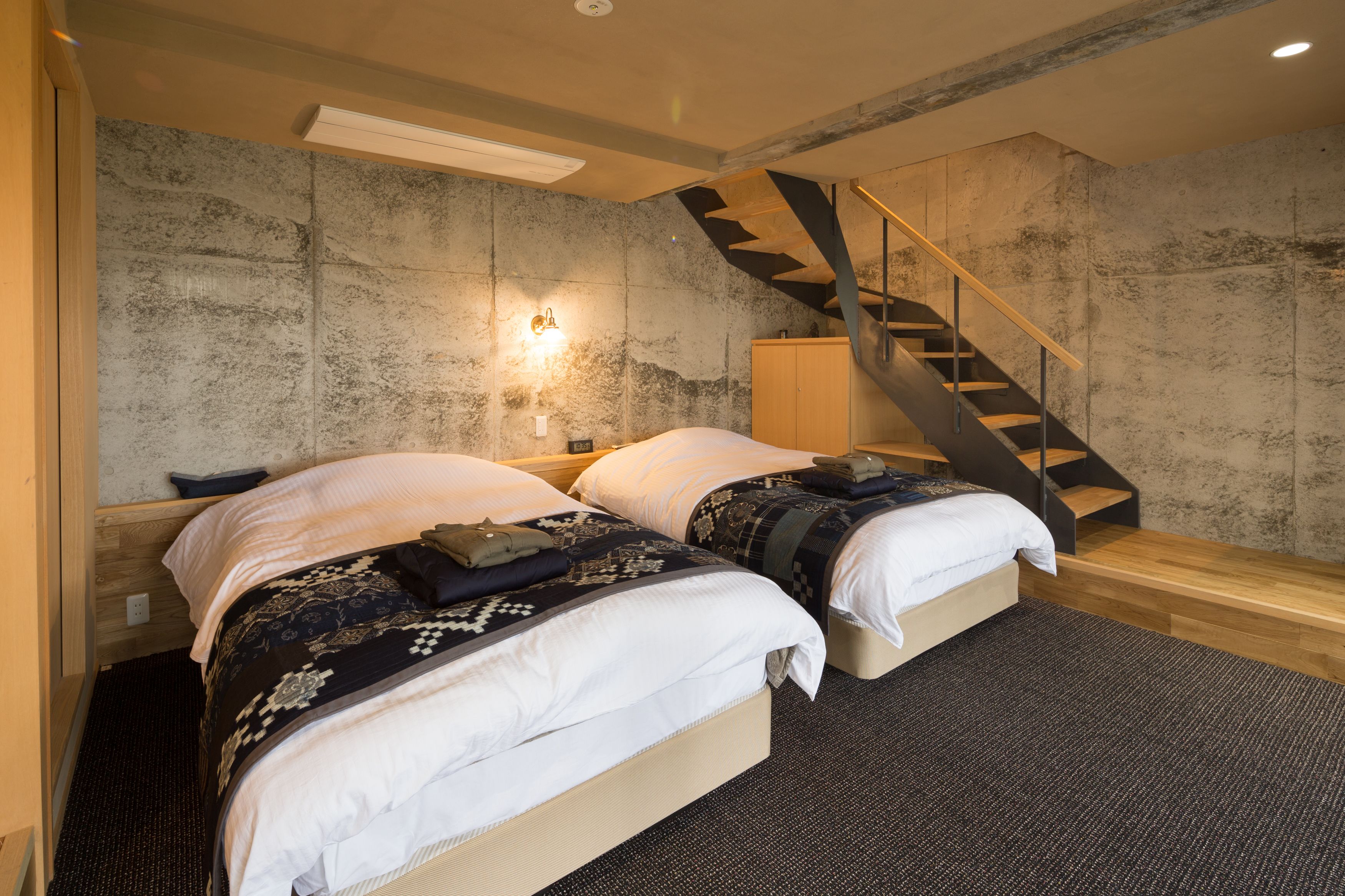 A modern bedroom with two beds, a wooden staircase, and a textured wall.