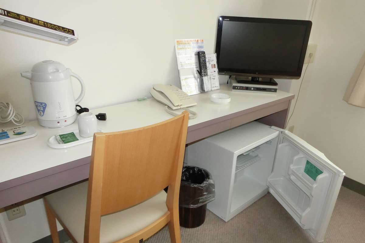 Habitación de hotel con escritorio, silla, cafetera, teléfono, televisor y cubo de basura.