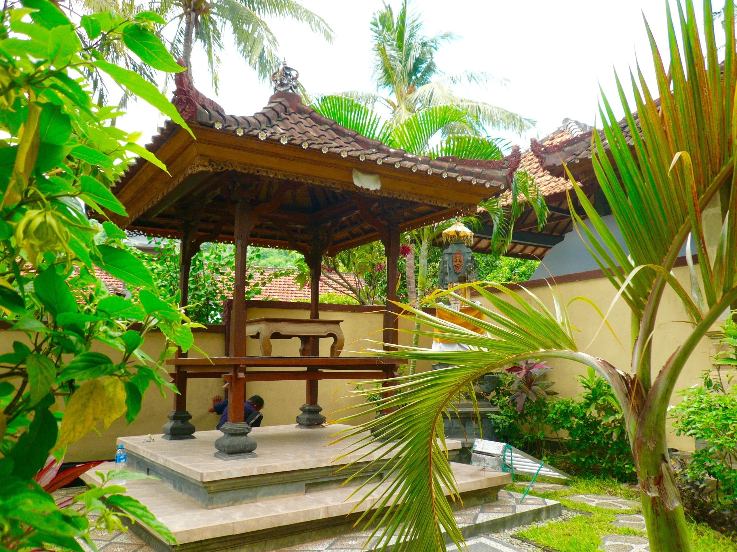 A traditional wooden gazebo with a tiled roof, surrounded by lush greenery and palm trees.