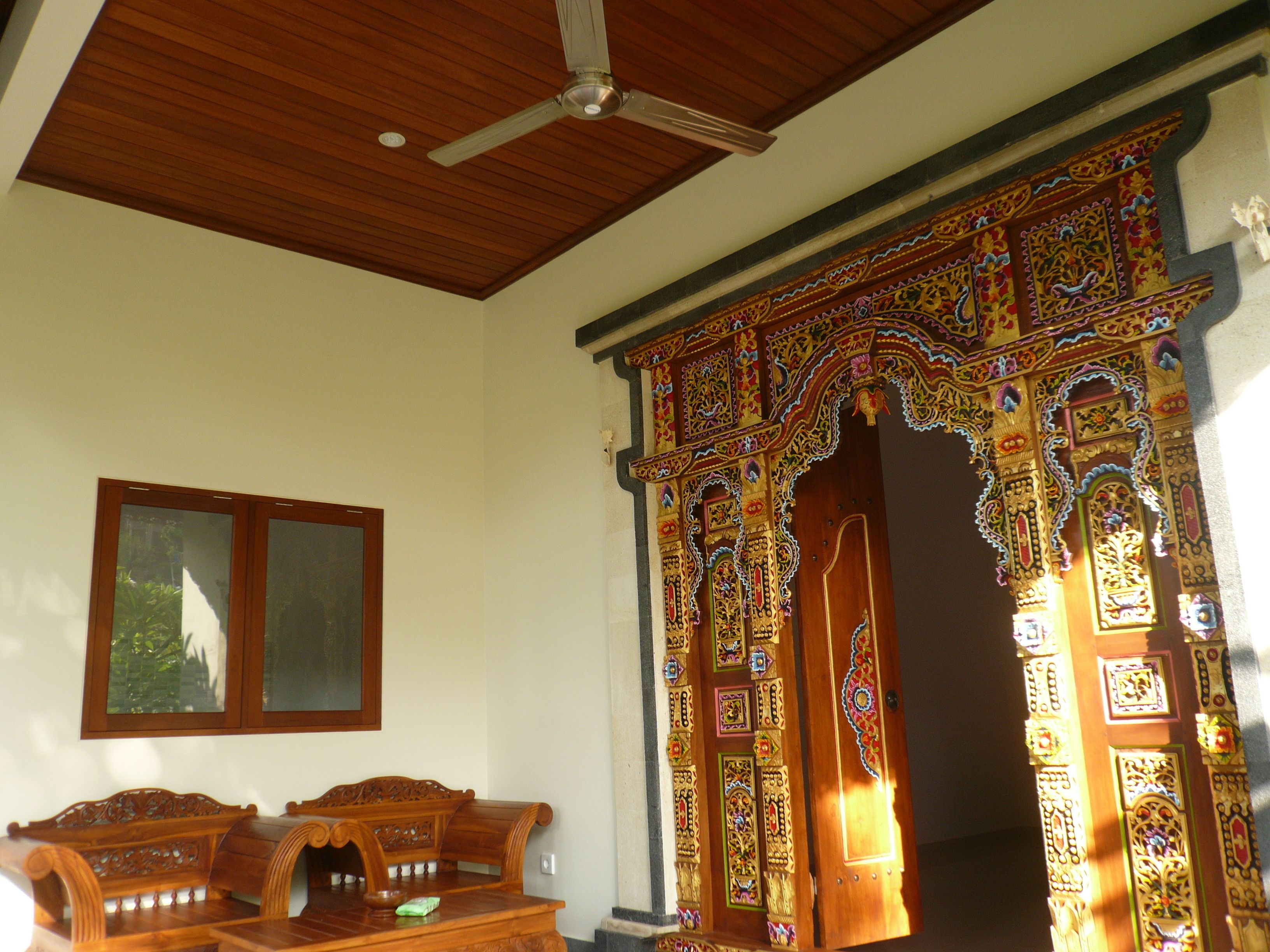 Een klassieke kamer met houten plafond, een plafondventilator, een houten bank en een decoratieve deur met ingewikkelde patronen.